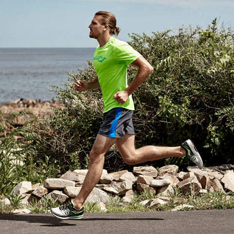 Eine Person in einem hellgrünen Hemd und blauen Shorts joggt auf einem gepflasterten Weg am Meer entlang, im Hintergrund sind Grünflächen und Felsen zu sehen.