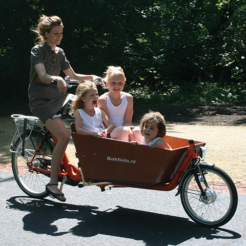 Eine Frau fährt an einem sonnigen Tag mit einem Lastenfahrrad. Drei Kinder sitzen vorne im Fahrradkorb. Sie lächeln und genießen die Fahrt, umgeben von viel Grün.