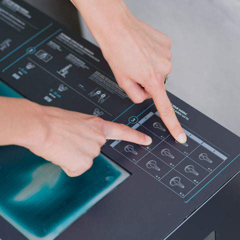 Zwei Hände interagieren mit einem Touchscreen-Panel. Der Bildschirm zeigt eine Auswahl von Symbolen, jedes davon zeigt ein Ohr mit Zahlen von 1 bis 10. Der Kontext schlägt vor, Audioeinstellungen oder Hörhilfeoptionen anzupassen.