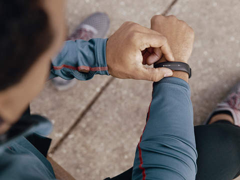 Eine Person in einem blauen Langarmshirt blickt auf einen Fitnesstracker an ihrem Handgelenk. Sie sitzt, ein Bein ist sichtbar, und trägt grau-rote Laufschuhe. Der Hintergrund ist eine Betonfläche.