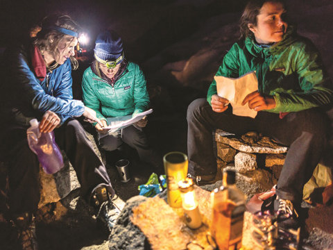 Drei Personen in Winterkleidung sitzen nachts auf einem Campingplatz. Im Licht ihrer Stirnlampen studieren sie Karten oder Papiere. Auf einem kleinen provisorischen Tisch stehen Kerzen, Flaschen und Tassen. Die Umgebung ist rau und im Freien.