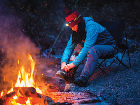 Eine Person in blauer Jacke und roter Mütze sitzt mit einer Stirnlampe auf dem Kopf nachts auf einem Campingstuhl neben einem Lagerfeuer und rückt ihre Wanderschuhe zurecht. Das Feuer wirft einen warmen Schein, der einen Kontrast zur dunklen Umgebung bildet.