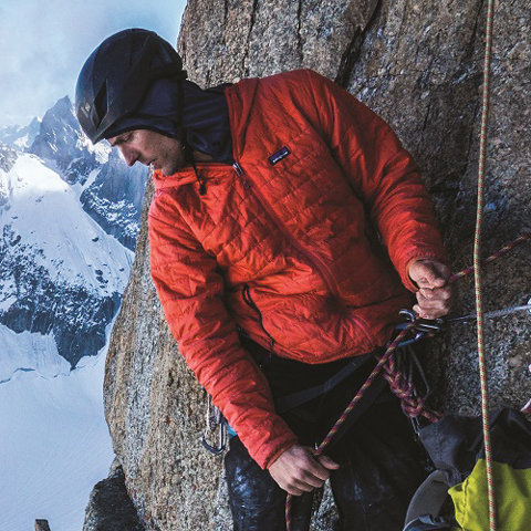 Ein Kletterer in roter Jacke und Helm befestigt Seile an einer felsigen Bergwand. Im Hintergrund sind schneebedeckte Gipfel unter einem bewölkten Himmel zu sehen. Die Szene vermittelt ein Gefühl von Abenteuer und Erkundung im Freien.