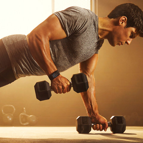 Ein Mann in einem grauen Hemd macht in einem Fitnessstudio Plank Row mit Hanteln und zeigt dabei definierte Muskeln. Sonnenlicht fällt durch ein Fenster und hebt sein Training hervor. Kettlebells sind im Hintergrund verschwommen.