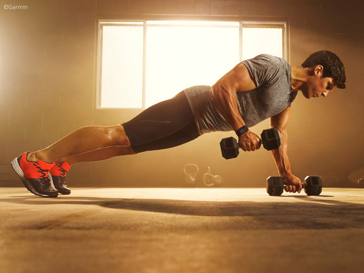 Eine Person in Sportkleidung führt in einem sonnendurchfluteten Fitnessstudio Plank Row mit Hanteln aus. Sie trägt rote und schwarze Schuhe und konzentriert sich auf Kraft und Gleichgewicht. Im Hintergrund sind ein großes Fenster und Kugelhanteln auf dem Boden zu sehen.