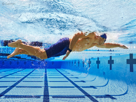 Unterwasseransicht eines Schwimmers in Bewegung in einem klaren Becken. Der Sportler trägt eine Badekappe, eine Schwimmbrille und eine Badehose und hat die Arme für einen Freistilzug ausgestreckt. Der Boden und die Wände des Beckens sind mit blauen Fliesen bedeckt, im Hintergrund sind schwarze Pluszeichen zu sehen.