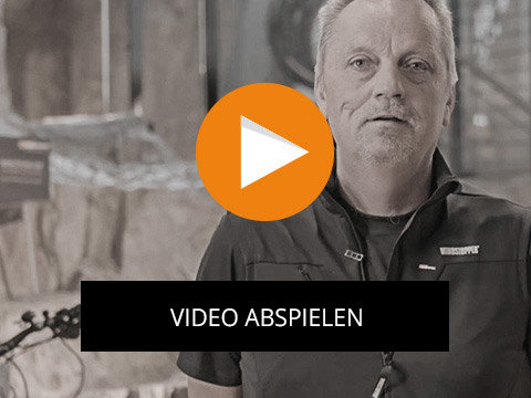 Ein Mann in einem dunklen Hemd steht im Innenbereich, im Hintergrund ist ein Fahrrad zu sehen. Über dem Bild ist ein orangefarbener Play-Button mit dem Text „VIDEO ABSPIELEN“ eingeblendet.