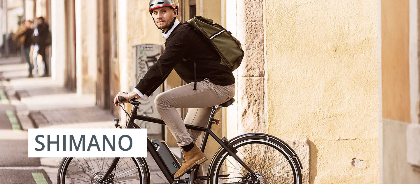 Ein Mann mit Helm und Rucksack fährt mit dem Fahrrad eine von Gebäuden gesäumte Straße entlang. Links ist das Wort „Shimano“ zu sehen.