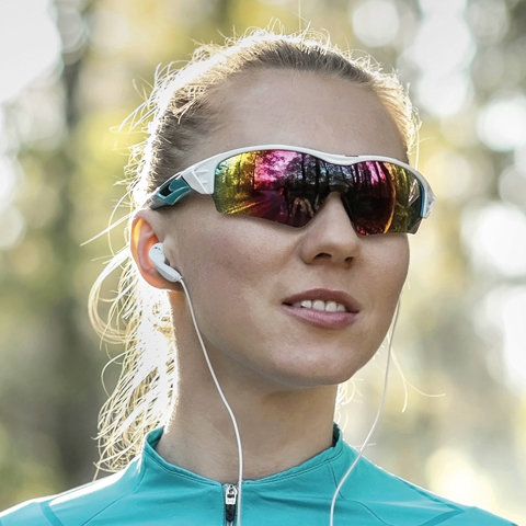 Eine Frau mit verspiegelter Sonnenbrille und Ohrstöpseln lächelt im Freien. Sie trägt eine türkisfarbene Sportjacke und im Hintergrund ist eine verschwommene Naturlandschaft mit Sonnenlicht zu sehen, das durch die Bäume fällt.