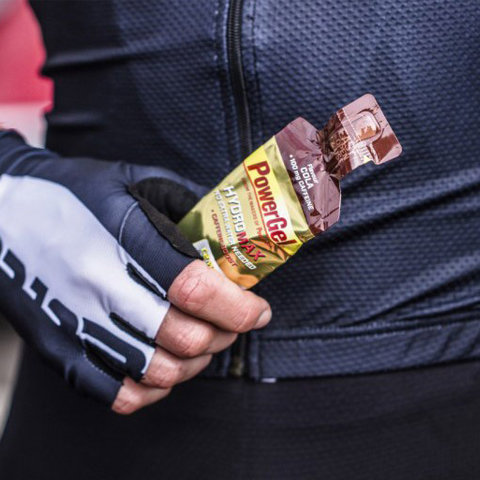 Nahaufnahme einer Person in schwarzer Sportkleidung und Handschuhen, die ein PowerGel-Energiegelpack in der Hand hält. Das Gelpack ist gelb und braun, was auf eine Geschmacksvariante hindeutet. Die andere Hand der Person ist teilweise sichtbar.