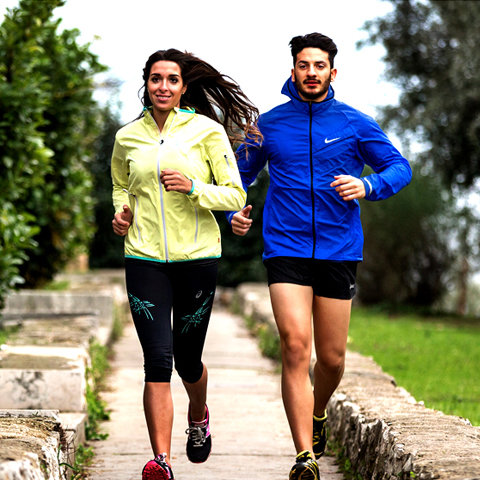 Eine Frau in einer gelben Jacke und ein Mann in einer blauen Jacke joggen nebeneinander auf einem Steinweg. Sie sind im Freien, umgeben von Grün, und bewegen sich auf die Kamera zu. Beide tragen Sportschuhe.