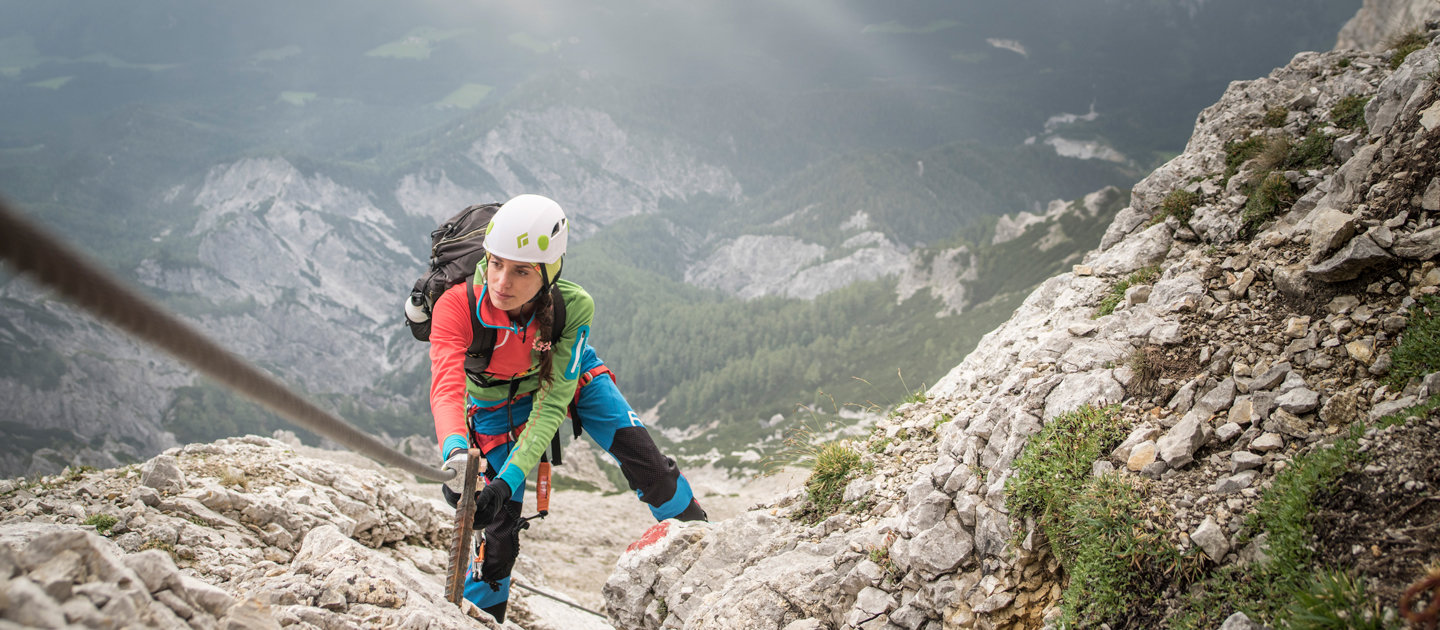 Ein Bergsteiger in farbenfroher Ausrüstung und einem weißen Helm erklimmt mithilfe eines gesicherten Seils einen steilen, felsigen Berghang. Der Hintergrund zeigt eine verschwommene Landschaft mit Wäldern und fernen Gipfeln unter einem bewölkten Himmel.