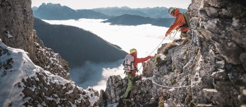 Zwei Kletterer mit Helmen und Ausrüstung erklimmen einen felsigen Berg, umgeben von Wolken und Gipfeln in der Ferne. Ein Kletterer hilft dem anderen mit einem Seil, während sie durch das zerklüftete Gelände navigieren.