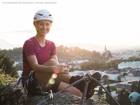 Eine Person in Kletterausrüstung, mit Helm und magentafarbenem Hemd, sitzt auf einer felsigen Oberfläche und genießt eine malerische Aussicht. Im Hintergrund sind eine sonnenbeschienene Stadt und Hügel in der Ferne zu sehen.