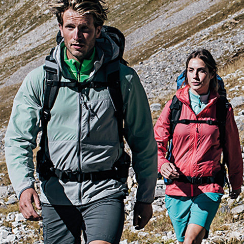 Zwei Personen wandern in einer bergigen Gegend. Der Mann im Vordergrund trägt eine graue Jacke und Shorts, während die Frau im Hintergrund eine rote Jacke und blaue Shorts trägt. Beide haben Rucksäcke dabei und bewegen sich über felsiges Gelände.