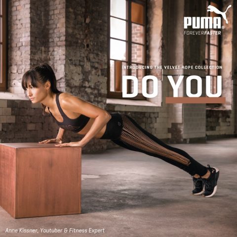 Eine Frau macht in einem Raum im Industriestil eine Plank-Übung mit den Händen auf einem Holzblock. Sie trägt ein schwarzes Sportoutfit. Der Text auf dem Bild enthält „Puma“, „Wir stellen die Velvet Rope Collection vor“ und „Do You“.