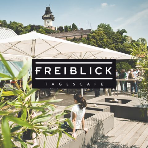 Ein Café auf dem Dach namens Freiblick Tagescafé mit Menschen, die das sonnige Wetter genießen. Es bietet große Sonnenschirme, Sitzgelegenheiten und Grünflächen. Im Hintergrund ist vor einem klaren Himmel ein Glockenturm zu sehen.