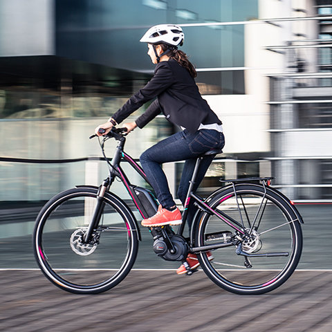 Eine Person mit Helm und Blazer fährt in einer städtischen Umgebung auf einem schwarz-rosa Fahrrad. Der Radfahrer ist in Bewegung und der Hintergrund ist verschwommen, was die Geschwindigkeit hervorhebt. Sie trägt Jeans und rote Turnschuhe.