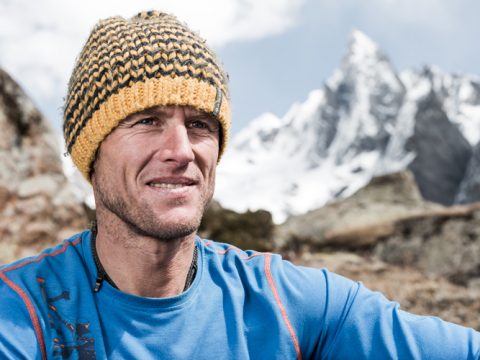 Eine Person mit einer gelb-schwarzen Strickmütze und einer blauen Jacke steht im Freien. Im Hintergrund sind schneebedeckte Berge unter einem bewölkten Himmel zu sehen. Die Person blickt zur Seite und scheint sich in einer Bergregion zu befinden.