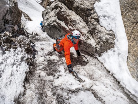 Ein Bergsteiger in orangefarbener Jacke und Helm erklimmt mit Eispickeln eine steile, eisige Felswand. Das anspruchsvolle Gelände ist von Schnee und Felsen umgeben. Der Bergsteiger wirkt konzentriert und entschlossen.