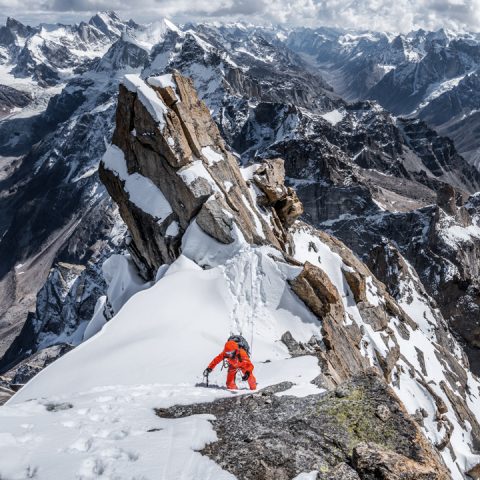 Ein Bergsteiger in roter Montur erklimmt einen schneebedeckten, zerklüfteten Bergkamm mit steilen Felsformationen im Hintergrund. Schneebedeckte Gipfel erstrecken sich in der Ferne unter einem wolkigen Himmel.