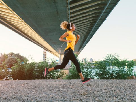 Eine Person in einem gelben Shirt und schwarzen Leggings joggt unter einer Brücke. Sie hört Musik über Kopfhörer und läuft auf einem Schotterweg. Im Hintergrund sind Grünflächen und ein Gewässer zu sehen.