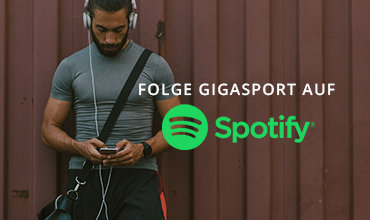 Ein Mann mit Kopfhörern und sportlichem Outfit schaut auf sein Handy. Der Text lautet „FOLGE GIGASPORT AUF Spotify“. Ein Spotify-Logo wird angezeigt. Der Hintergrund ist eine braune Wand.
