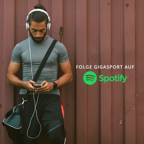 Ein Mann mit Kopfhörern lehnt an einer braunen Wand und schaut auf sein Handy. Er trägt ein graues Hemd, schwarze Shorts und hat eine Tasche über der Schulter. Darauf steht der Text „Folge Gigasport auf Spotify“ mit dem Spotify-Logo.
