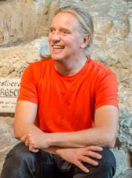 Lächelnder Mann mit langen Haaren und orangefarbenem Hemd, der mit verschränkten Armen vor einem rauen Steinhintergrund mit einem teilweise sichtbaren Schild sitzt.