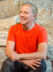 Lächelnder Mann mit langen Haaren und orangefarbenem Hemd, der mit verschränkten Armen vor einem rauen Steinhintergrund mit einem teilweise sichtbaren Schild sitzt.