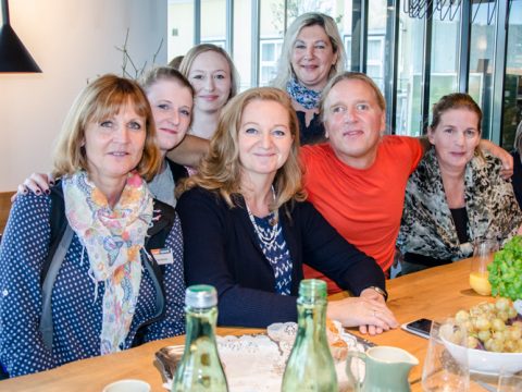 Eine Gruppe von sieben Personen sitzt und steht um einen Holztisch herum und lächelt in die Kamera. Auf dem Tisch stehen Flaschen, ein Teller mit Deckchen und eine Schale mit Weintrauben. Im Hintergrund sind Fenster und Inneneinrichtung zu sehen.