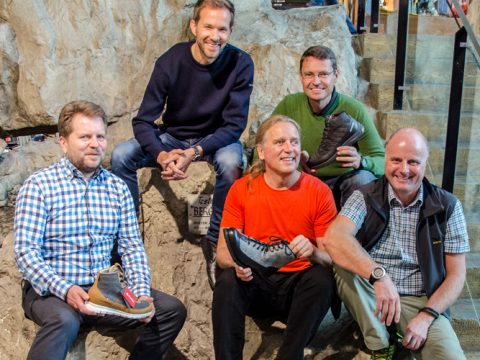 Fünf Männer sitzen und stehen auf Felsstufen, jeder hält Wanderschuhe in einer Verkaufsumgebung. Sie scheinen zu lächeln und leger gekleidet zu sein. Im Hintergrund ist eine Steinmauer zu sehen.