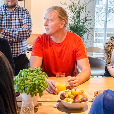 Eine Person in einem roten Hemd sitzt mit einem Glas Orangensaft an einem Tisch. Eine Schale mit Obst und eine Basilikumpflanze stehen auf dem Tisch. Andere Personen sind teilweise um den Tisch herum zu sehen. Die Atmosphäre wirkt leger und drinnen.