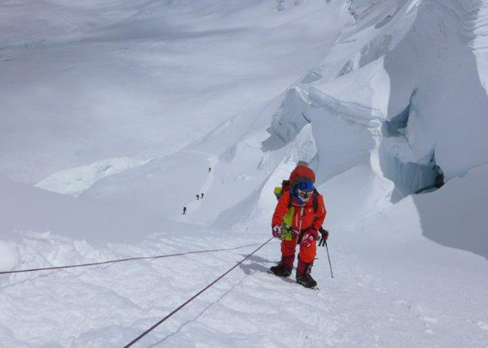 Ein Bergsteiger in roter Winterkleidung erklimmt einen schneebedeckten Hang und hält sich dabei an einem Seil fest. In der Ferne sind mehrere andere Bergsteiger zu sehen, die einen ähnlichen Weg entlanggehen. Die Landschaft ist schneebedeckt und schroff, was auf eine Bergbesteigung in großer Höhe schließen lässt.