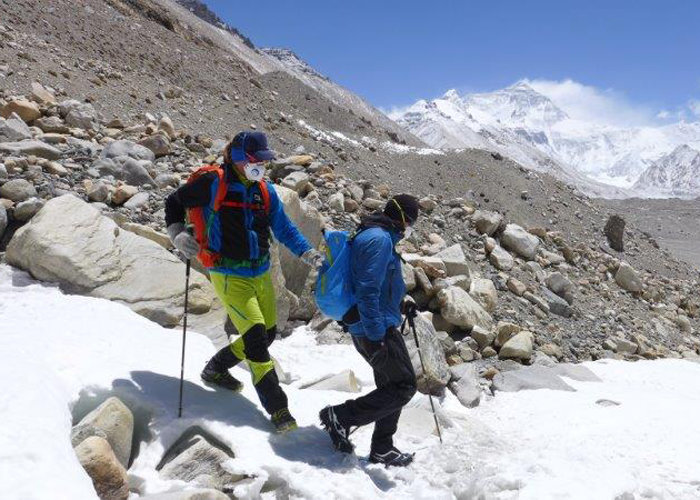 Zwei Bergsteiger überqueren einen schneebedeckten Berghang. Sie tragen farbenfrohe Ausrüstung und Masken. Sie benutzen Trekkingstöcke zur Stabilisierung. Im Hintergrund sind das felsige Gelände und die schneebedeckten Gipfel unter einem klaren blauen Himmel zu sehen.