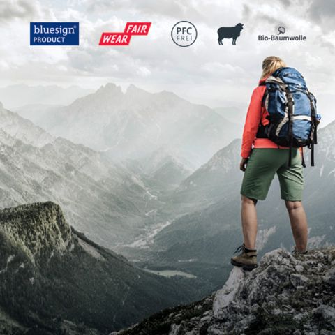 Ein Wanderer steht auf einem Berggipfel und überblickt eine weite Landschaft aus Tälern und Bergen. Symbole oben weisen auf Bluesign-Produkte, Fair Wear, PFC-freie Produkte und Bio-Baumwolle hin. Die Person trägt Wanderausrüstung und einen großen Rucksack.