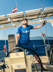 Ein Mann in blauer Sportkleidung steuert ein Segelboot auf offenem Wasser unter einem klaren blauen Himmel. Er hält mit einer Hand ein Seil und steht hinter dem Steuerrad des Schiffs, mit Blick in den Himmel. Im Hintergrund weht eine Flagge.