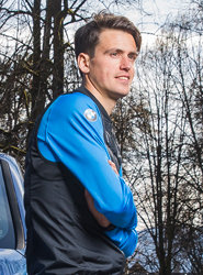 Ein Mann mit kurzen braunen Haaren und einer blau-schwarzen Jacke steht mit verschränkten Armen im Freien. Im Hintergrund sind Bäume und ein bewölkter Himmel zu sehen.