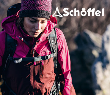 Eine Person mit rot-rosa Outdoorjacke und Strickmütze wandert mit Rucksack. Im Hintergrund sind Felsen und Bäume unscharf zu erkennen. Oben rechts ist das Schöffel-Logo zu sehen.