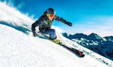 Ein Skifahrer in dunkler Kleidung, mit Helm und Schutzbrille, fährt einen schneebedeckten Abhang hinunter. Hinter ihnen spritzt der Schnee, als sie scharf abbiegen. Der Himmel ist klar und blau, im Hintergrund sind schneebedeckte Berge zu sehen.
