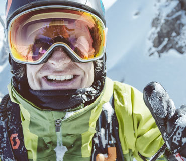 Eine Person in grüner Jacke, Skibrille und Helm lächelt und zeigt mit dem Daumen nach oben. Im Hintergrund sind schneebedeckte Berge zu sehen, die auf ein Ski- oder Snowboard-Abenteuer hindeuten.
