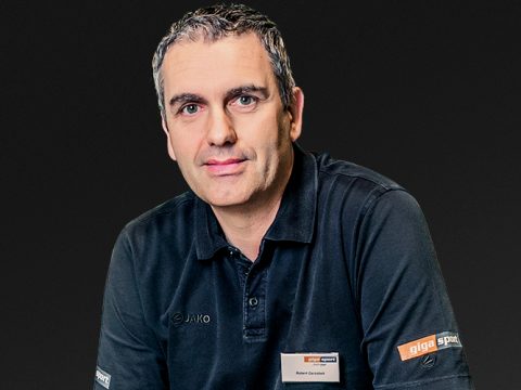 Mann mit kurzen grauen Haaren, trägt ein schwarzes Jako-Poloshirt und ein Badge mit „gigasport“-Logo und posiert vor einem dunklen Hintergrund.