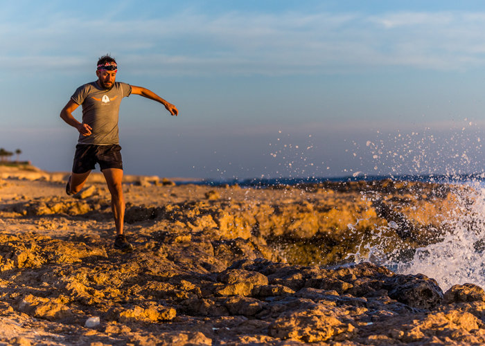 Ein Mann in Sportkleidung läuft an einer felsigen Küste entlang. Wellen schlagen gegen die Felsen, während die Sonne untergeht und die Szenerie in einen warmen Glanz taucht. Der Himmel ist teilweise bewölkt und weist Schattierungen von Blau und Orange auf.