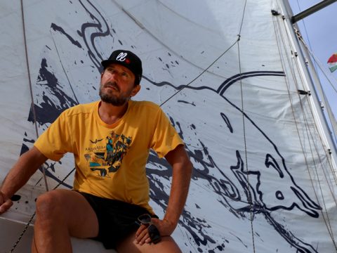 Ein Mann mit gelbem Hemd und schwarzer Mütze sitzt auf einem Segelboot. Hinter ihm ist auf dem Segel die schwarzweiße Abbildung eines großen Tieres zu sehen. Am Mast ist eine Flagge zu sehen.