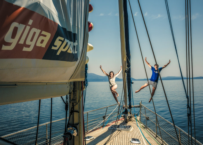 Zwei Menschen springen freudig vom Deck eines Segelboots ins ruhige Meer. Im Vordergrund ist das Segel mit der Aufschrift „Giga Sport“ zu sehen. Der Himmel ist klar und am Horizont sind in der Ferne Berge zu sehen.