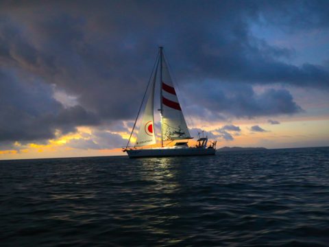 Ein Segelboot mit einem rot-weißen Segel wird bei Sonnenuntergang auf einem dunklen Meer beleuchtet. Der Himmel ist voller dunkler Wolken und in der Nähe des Horizonts befindet sich ein Streifen gelb-orangefarbenen Lichts.