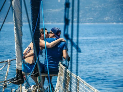 Ein Paar umarmt und küsst sich auf dem Deck eines Segelboots, umgeben von ruhigem blauen Wasser. Sie sind beide blau gekleidet, und der Mann trägt eine Mütze. Der sonnige Tag unterstreicht die ruhige und romantische Umgebung.