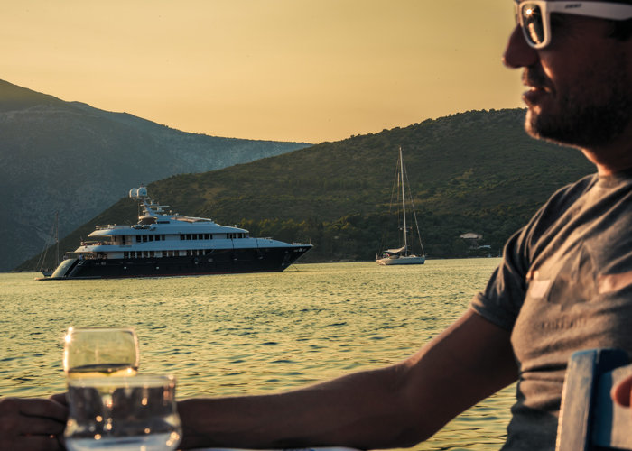 Eine Person mit Sonnenbrille sitzt mit einem Drink da und blickt auf das ruhige Meer bei Sonnenuntergang. Im Hintergrund ankern eine große Yacht und ein kleineres Segelboot in der Nähe hügeliger Küsten unter einem goldenen Himmel.