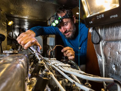 Eine Person mit Stirnlampe arbeitet in einem Metallraum an einem komplexen Maschinenaufbau. Sie benutzt ein Werkzeug, um Drähte und Komponenten unter hellem Licht anzupassen. In der Umgebung befinden sich verschiedene Kabel und eine 20-W-Leuchte.
