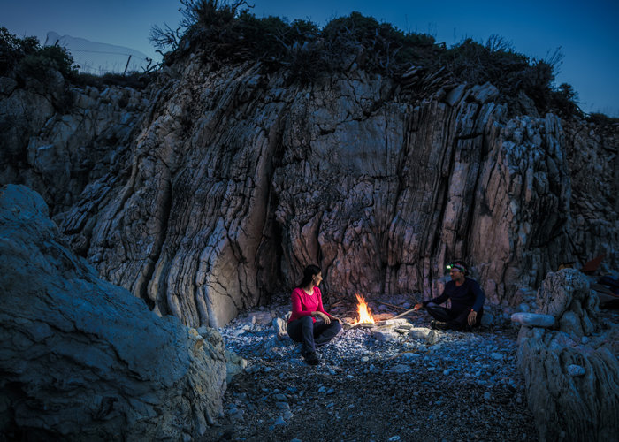 Zwei Personen sitzen in der Abenddämmerung an einem kleinen Lagerfeuer vor einer Felswand. Einer trägt eine Stirnlampe, der andere ein rotes Oberteil. Die Umgebung ist rau und weist steile, strukturierte Felsformationen auf, die eine ruhige Atmosphäre im Freien schaffen.